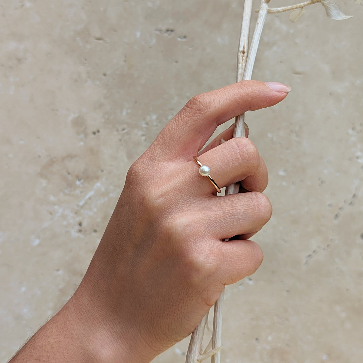 Hand holding a dried branch wearing a gold ring with a small round pearl